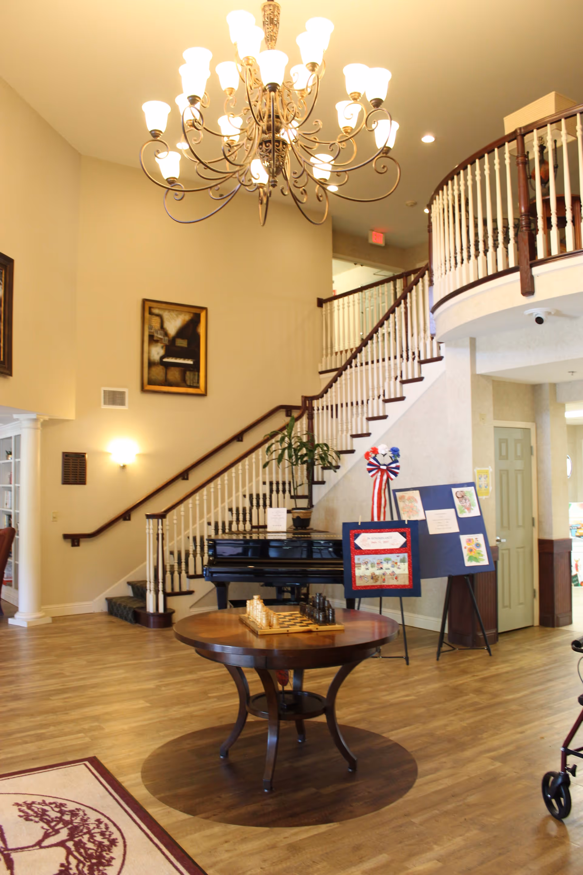 Interior view of a retirement community featuring a grand staircase with white railings and dark wood handrails, a large chandelier with multiple lights hanging from the ceiling, a round wooden table with a chess set on it, a black piano against the wall, and an easel displaying artwork and a red, white, and blue ribbon. The floor is wooden, and there is a patterned rug partially visible in the foreground.