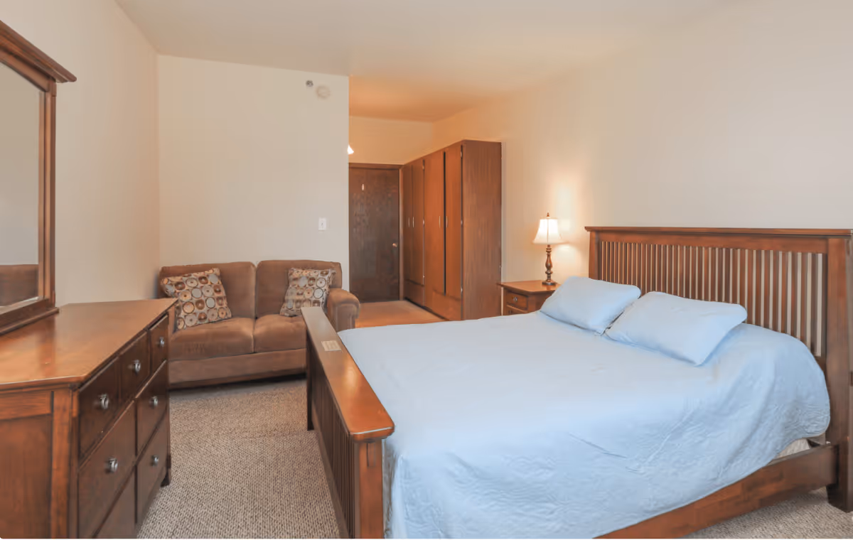 A bedroom in a senior living facility featuring a wooden bed with a light blue quilt and two pillows, a matching wooden dresser with a mirror, a brown loveseat with two patterned cushions, a wooden wardrobe, and a nightstand with a lit lamp against cream-colored walls.