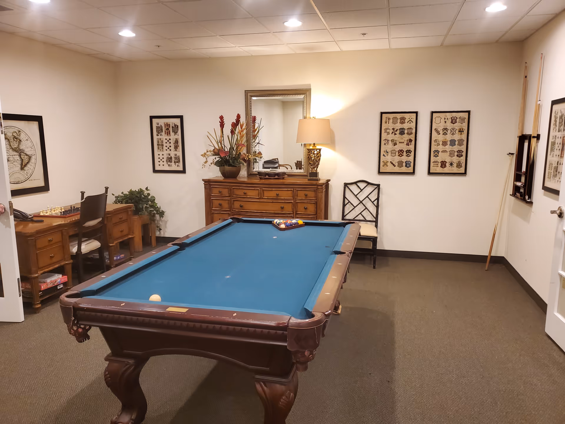 A recreational room with a pool table in the center, a wooden dresser with a lamp and flower arrangement against the back wall, framed artwork on the walls, a chair, and a desk with a chessboard and chair in the corner.