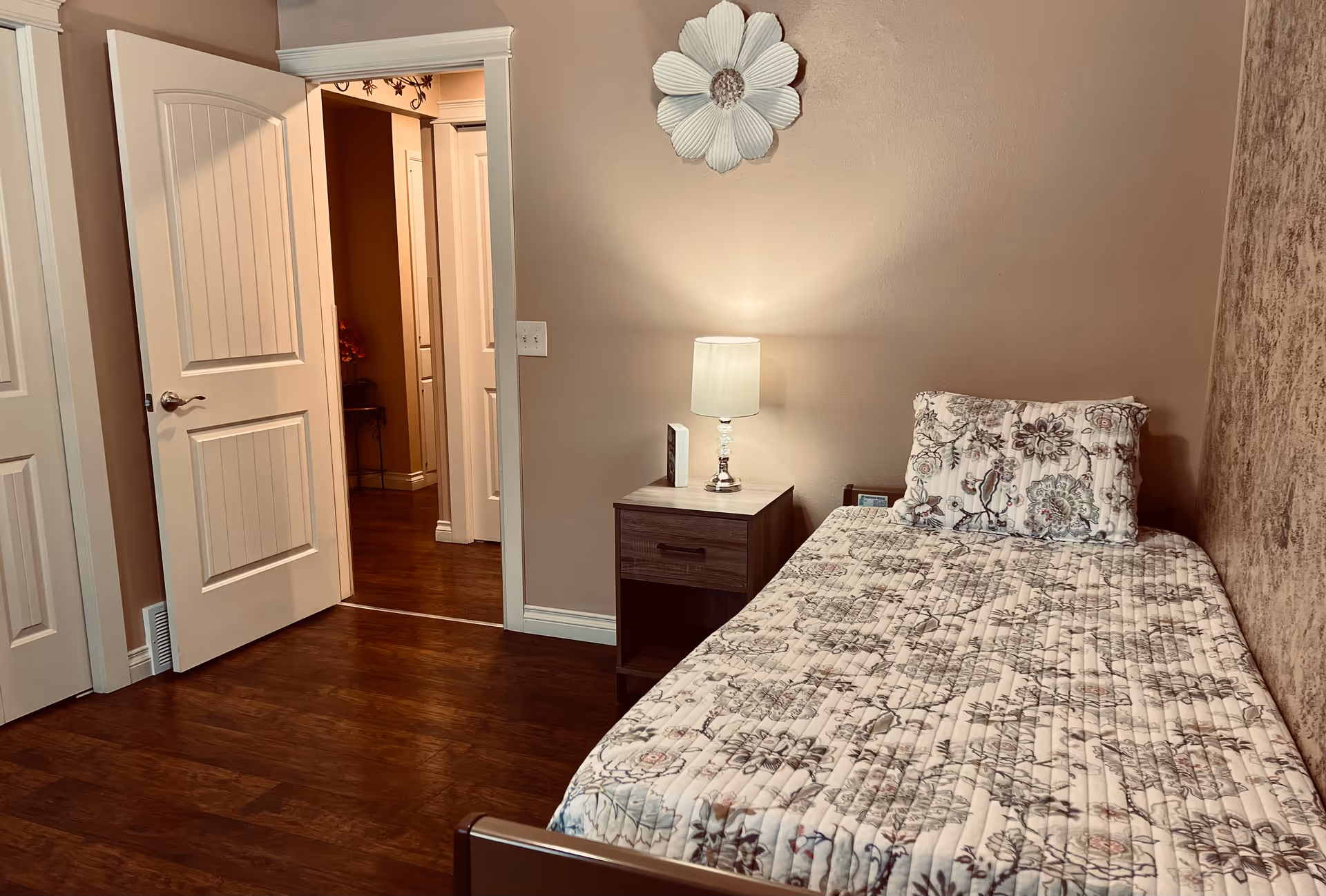 Small bedroom with a single bed dressed in floral bedding, a nightstand and lamp, and an open door to a hallway.