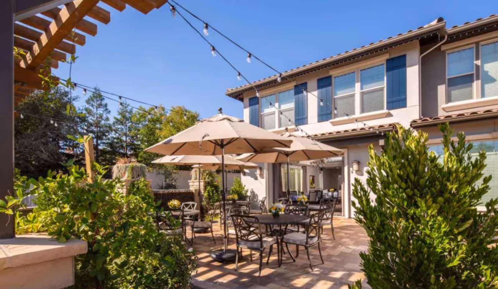 Outdoor patio area at Oakmont of Fair Oaks featuring multiple round tables with chairs and beige umbrellas. String lights are hung overhead, and there are green plants and trees surrounding the space. The building exterior with windows and blue shutters is visible in the background under a clear blue sky.