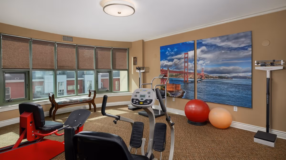 A small exercise room with various fitness equipment including a recumbent bike, an elliptical machine, and two large exercise balls. The room has a large window with brown blinds partially drawn, a wooden bench beneath the window, and a large two-panel wall art of the Golden Gate Bridge over water. The walls are beige and the floor is carpeted.
