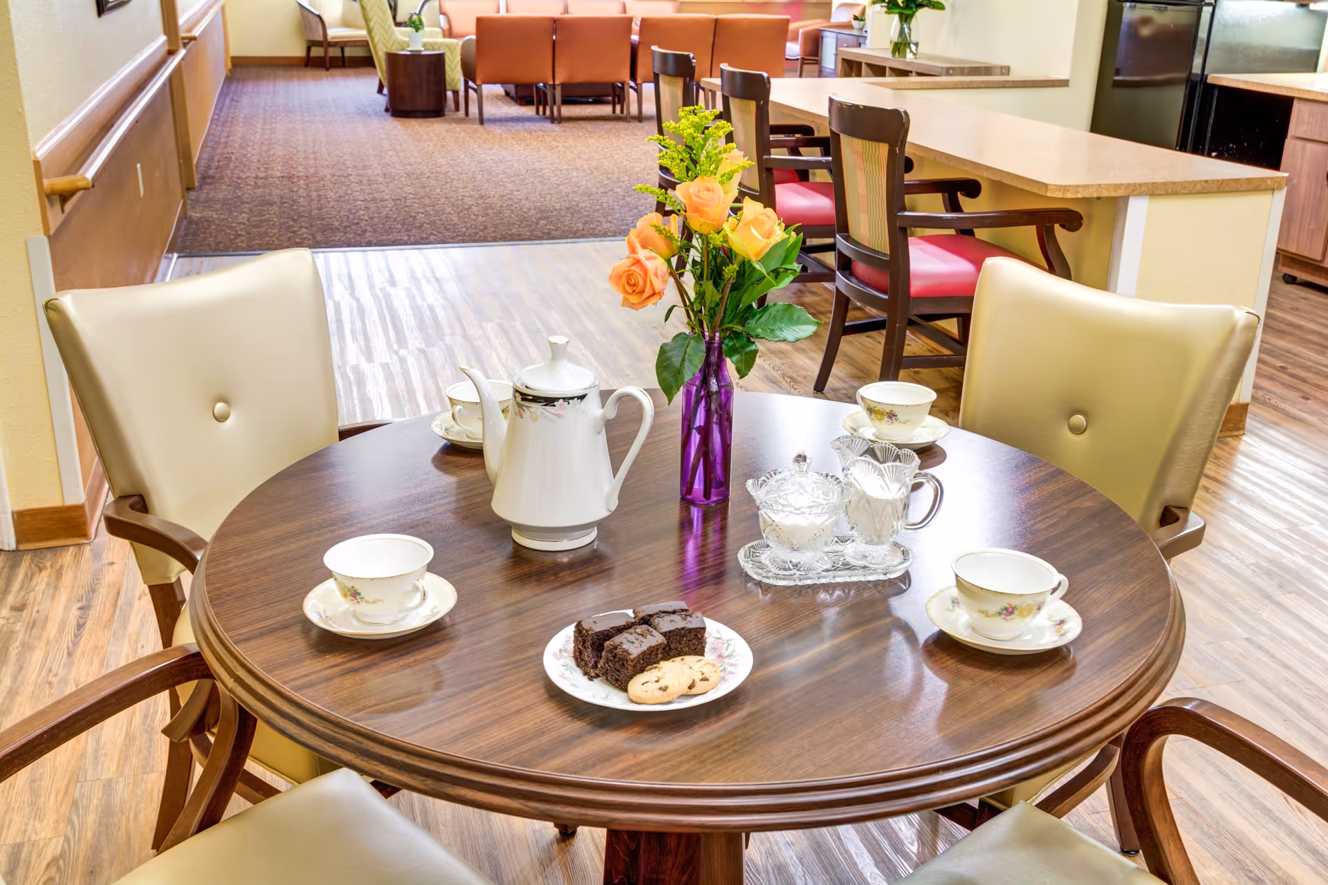 A round wooden table set for tea with a white teapot, four teacups and saucers, a plate with brownies and cookies, a sugar bowl, a creamer, and a purple vase with orange roses and greenery. The table is surrounded by four beige cushioned chairs. In the background, there are more chairs and tables in a well-lit room with wood and carpet flooring.
