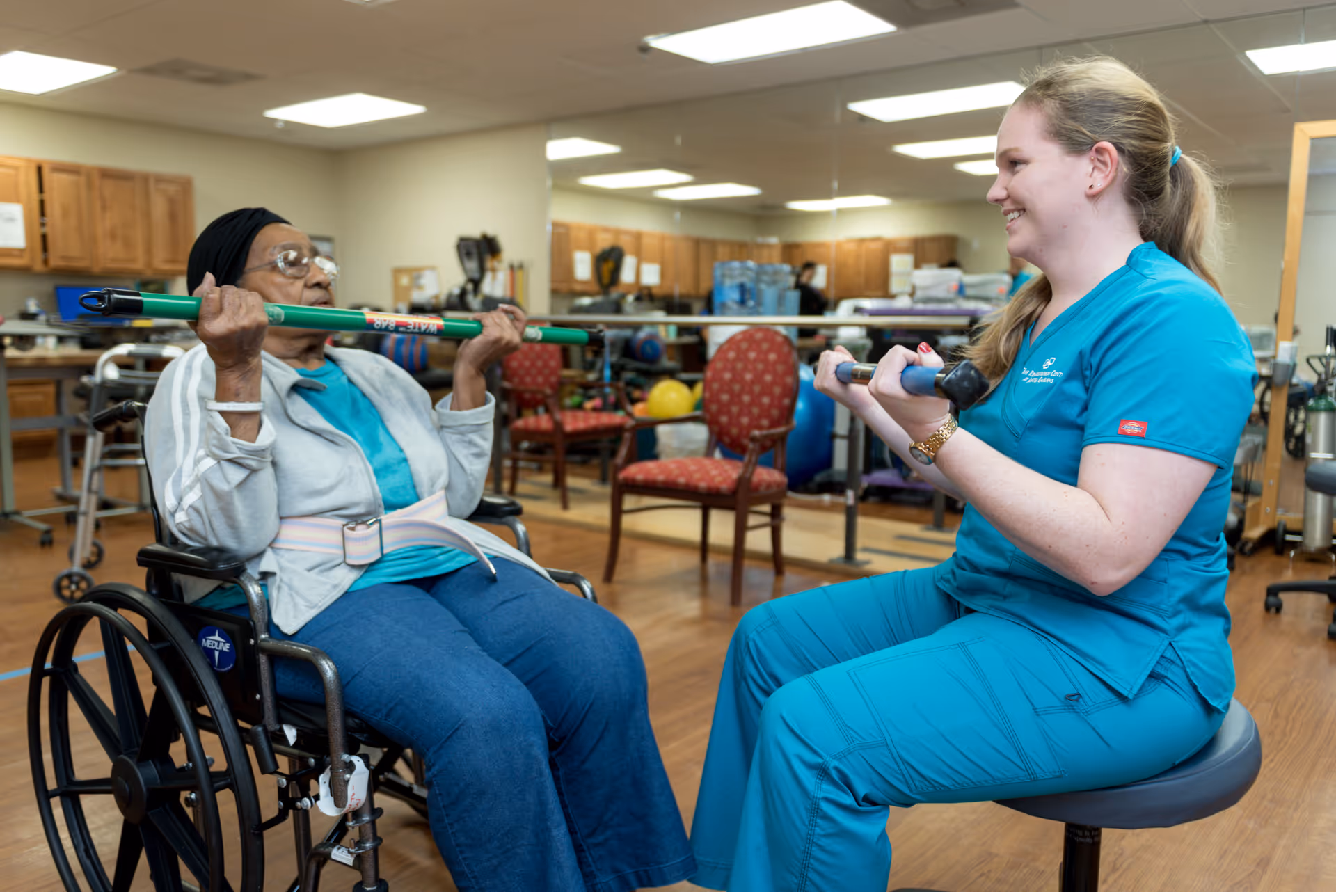 An elderly woman in a wheelchair is exercising with a green bar, assisted by a smiling female healthcare worker in blue scrubs seated on a stool in a rehabilitation center with exercise equipment and chairs in the background.