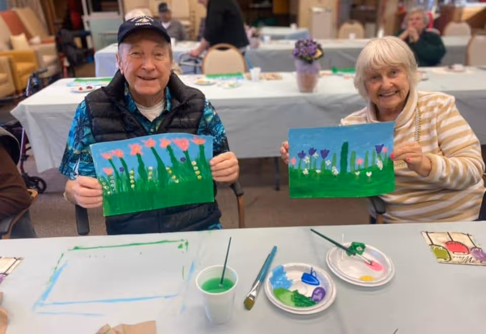 Two elderly individuals sitting at a table in a communal room, each holding up a colorful painting of flowers and greenery. The table in front of them has paintbrushes, paint palettes, and a cup with green paint. Other people and tables are visible in the background.