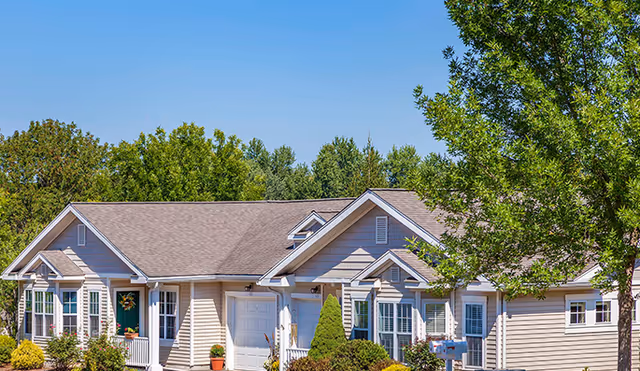 Exterior view of a single-story residential building with beige siding, white trim, and a brown shingled roof surrounded by green trees and shrubs under a clear blue sky.