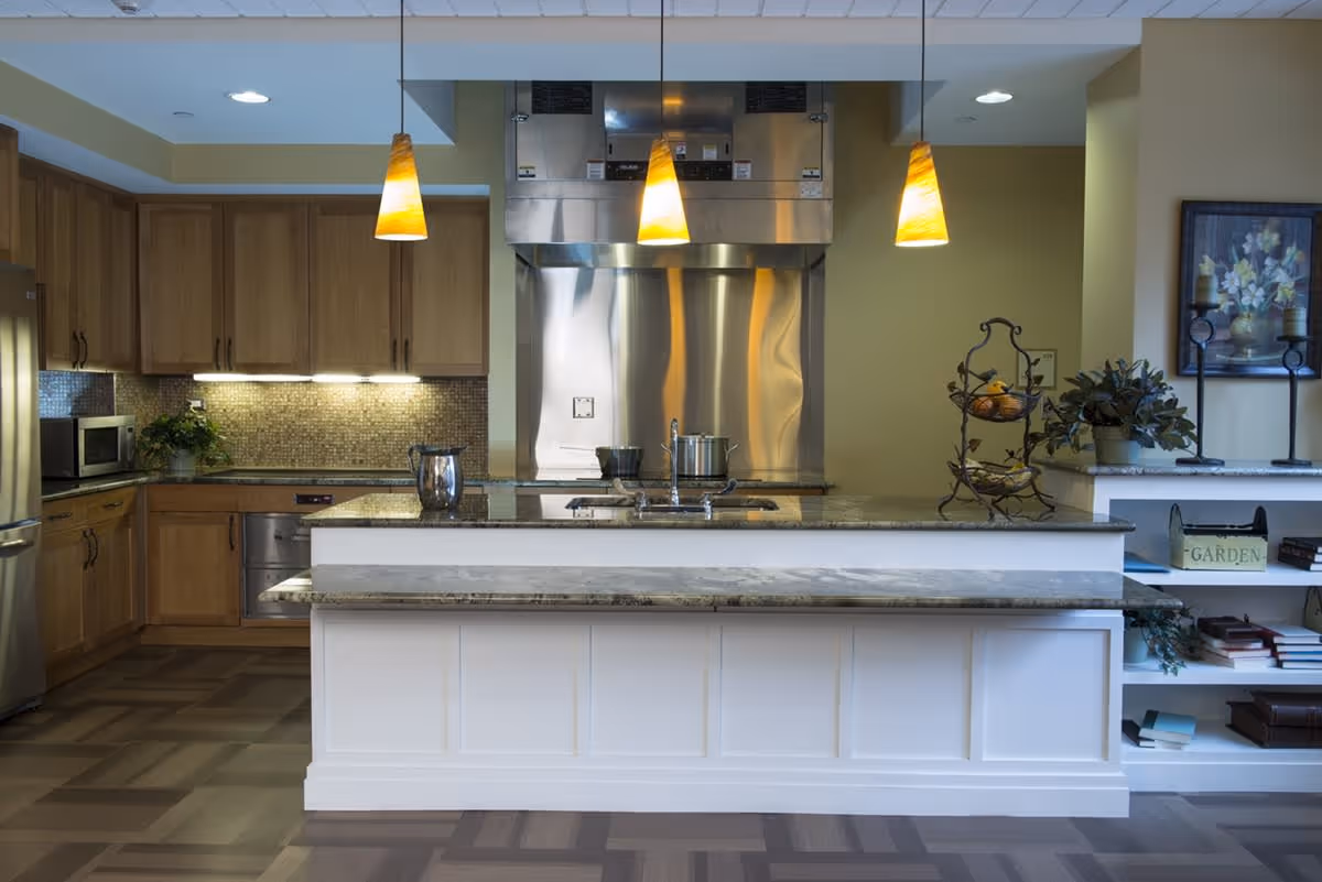 A modern kitchen with wooden cabinets, a stainless steel oven and microwave, a large island with a granite countertop, three hanging pendant lights, and decorative plants and items on shelves to the right.