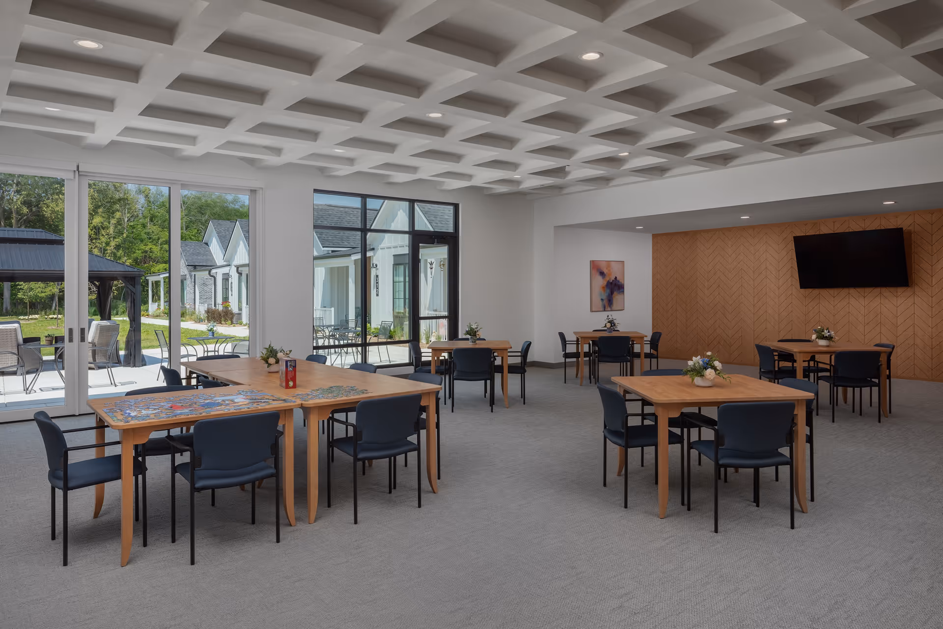 A spacious and well-lit common area with several wooden tables and black chairs arranged neatly. The room features large windows and glass doors that open to an outdoor patio with additional seating. The ceiling has a geometric pattern, and a flat-screen TV is mounted on a wooden accent wall. Small flower arrangements decorate the tables.