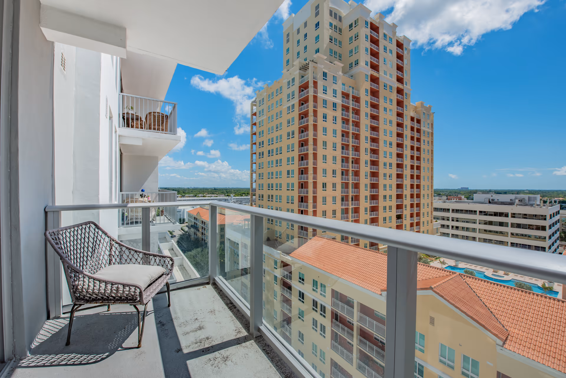 View from a balcony with a single wicker chair with a cushion, overlooking nearby buildings and a clear blue sky with some clouds.
