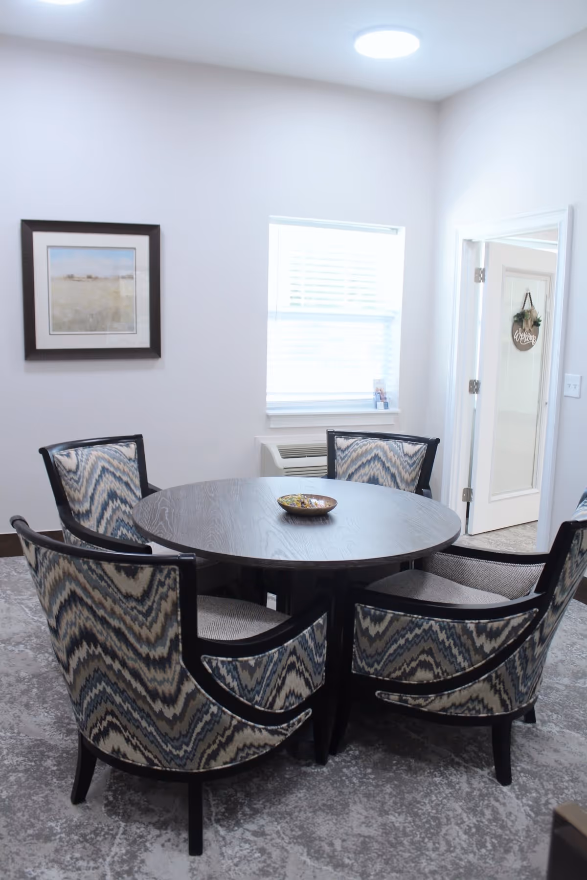 A small round wooden table surrounded by four upholstered chairs with a blue and beige zigzag pattern in a bright room with a window, a framed picture on the wall, and a door with a wreath hanging on it.