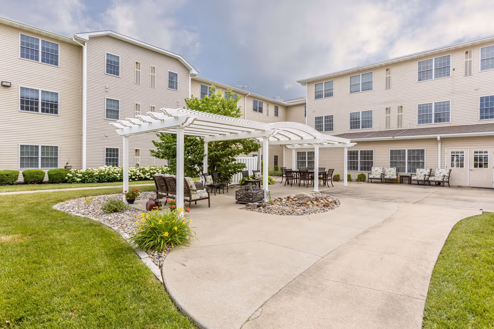 Outdoor courtyard with a white pergola, patio seating, and walkways in front of beige three-story residential buildings.