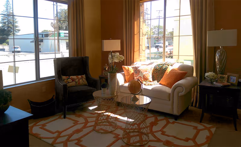 A cozy living room area with a white sofa adorned with orange and patterned throw pillows, a dark armchair with a decorative pillow, two round wireframe side tables with glass tops, a globe on one table, and two table lamps on either side. Large windows with beige curtains allow natural light to fill the room, and a patterned rug covers the floor.