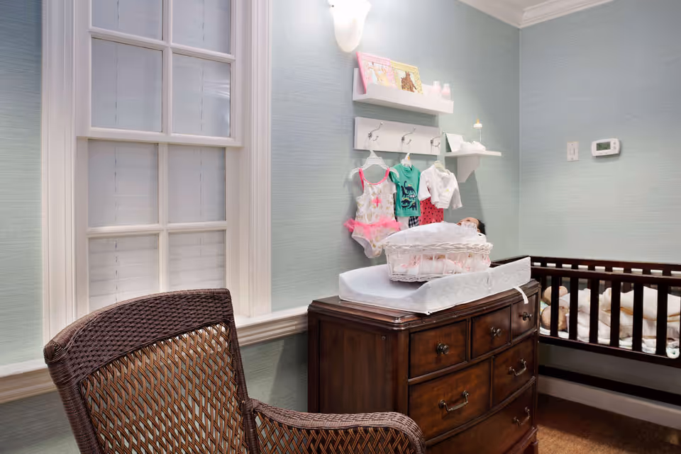 A nursery-style bedroom with a wicker chair, wooden dresser used as a changing table, hanging baby clothes on wall hooks, and a crib.
