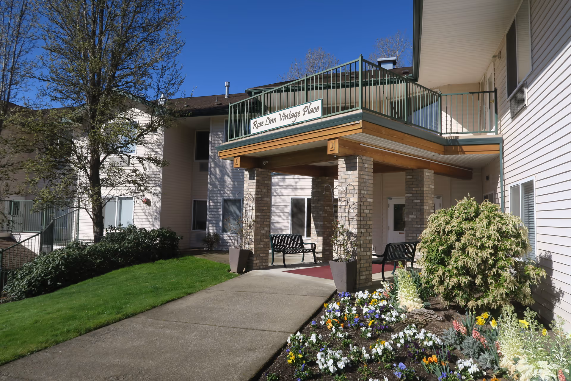 Entrance to Rose Linn Vintage Place, a senior living facility, featuring a covered porch supported by brick columns, benches, a walkway, green lawn, and a flower bed with colorful flowers under a clear blue sky.