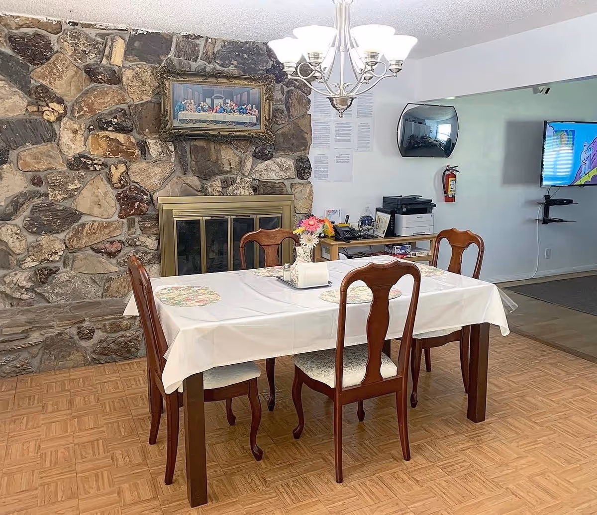 A dining area with a rectangular table covered with a white tablecloth and four wooden chairs. The table has floral placemats and a centerpiece with flowers and a napkin holder. Behind the table is a stone fireplace with a brass frame and a framed picture hanging above it. A chandelier with multiple lights hangs from the ceiling. In the background, there is a wall-mounted TV, a fire extinguisher, and a small desk with office equipment and board games.