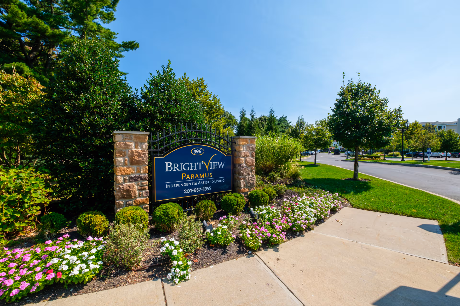 Outdoor view of the entrance sign for Brightview Paramus, a senior independent and assisted living facility, surrounded by green bushes, colorful flowers, and trees under a clear blue sky with a paved sidewalk and road nearby.