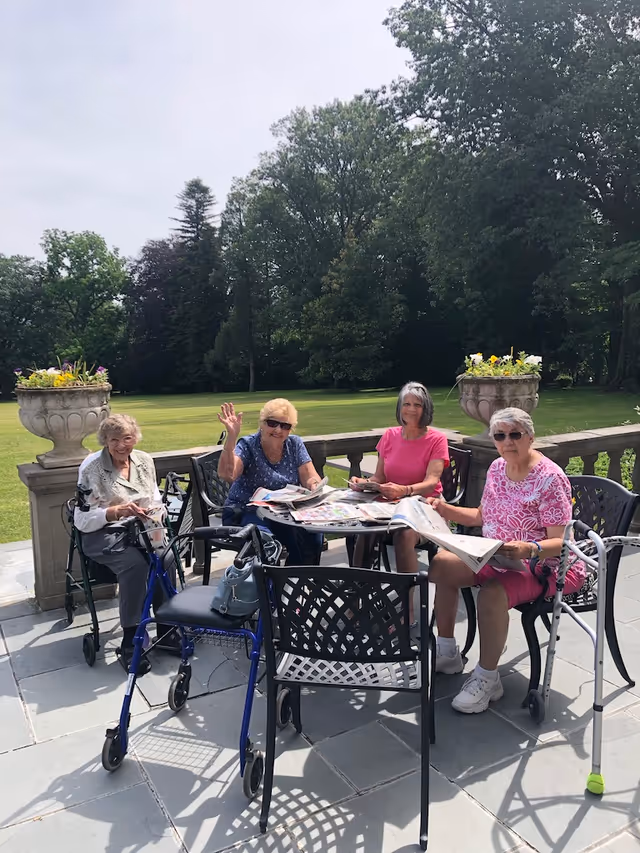 Four elderly women sitting around a black metal outdoor table on a stone patio, reading newspapers and smiling. Two women have walkers nearby. The background shows a large green lawn and tall trees under a partly cloudy sky.
