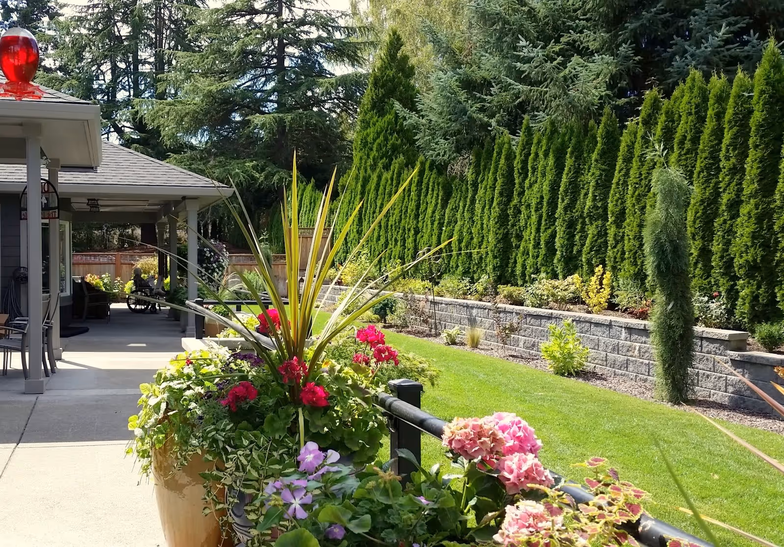 A well-maintained outdoor garden area at a senior care home featuring a variety of colorful flowers and plants in large pots along a paved walkway. There is a covered patio with seating and a red emergency light on the roof. Tall green trees and shrubs line the background, creating a serene and private environment.