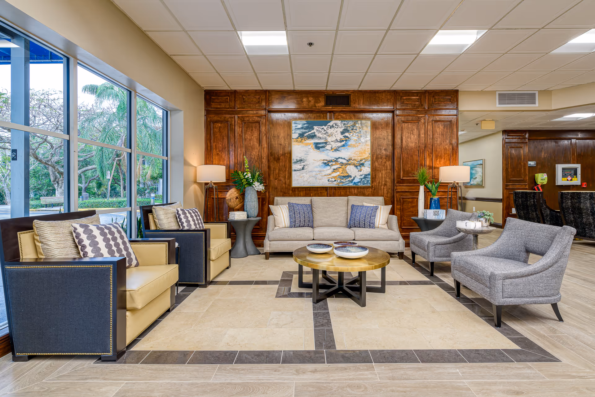 A bright senior living facility lobby with a sofa, armchairs, coffee tables, a wood-paneled wall, and large windows overlooking trees.