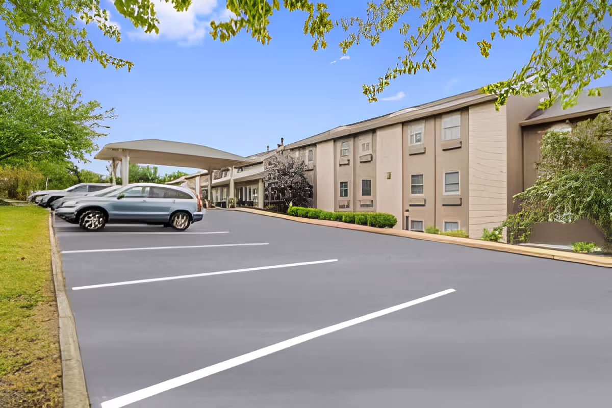 Exterior view of Rittenhouse Village At Muhlenberg showing a parking lot with several parked cars, a covered entrance, and a multi-story building surrounded by greenery under a clear blue sky.