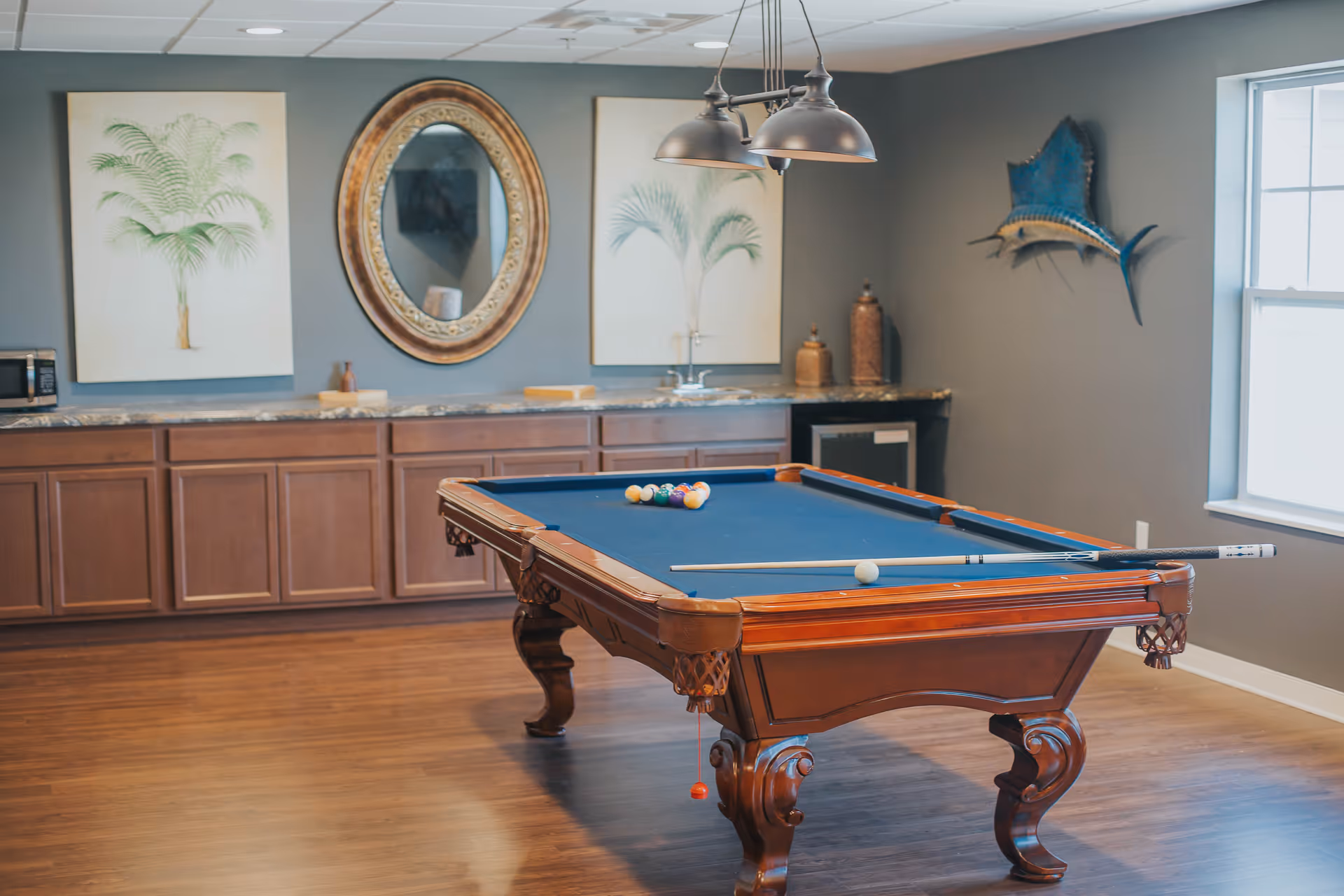 A recreational room with a wooden pool table featuring a blue felt surface, a cue stick, and billiard balls arranged for a game. The room has wooden flooring, a countertop with cabinets underneath, a microwave, a sink, decorative wall art including two framed palm prints, an ornate oval mirror, and a mounted fish sculpture. A window allows natural light into the space.
