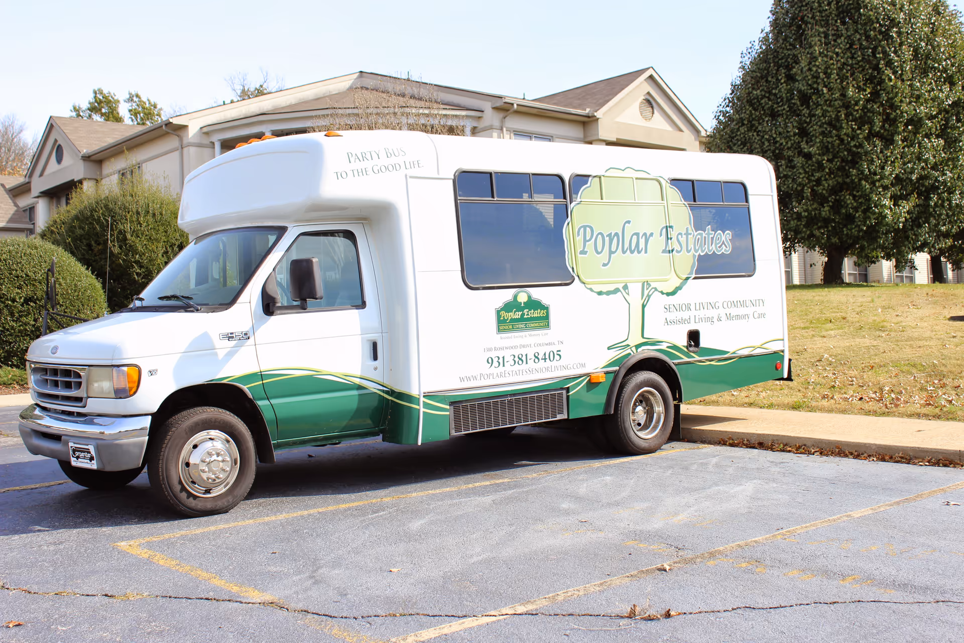 A white and green shuttle bus parked in a parking lot in front of a residential building. The bus has the logo and name 'Poplar Estates Senior Living Community' on the side, along with contact information and the slogan 'Party Bus to the Good Life.'