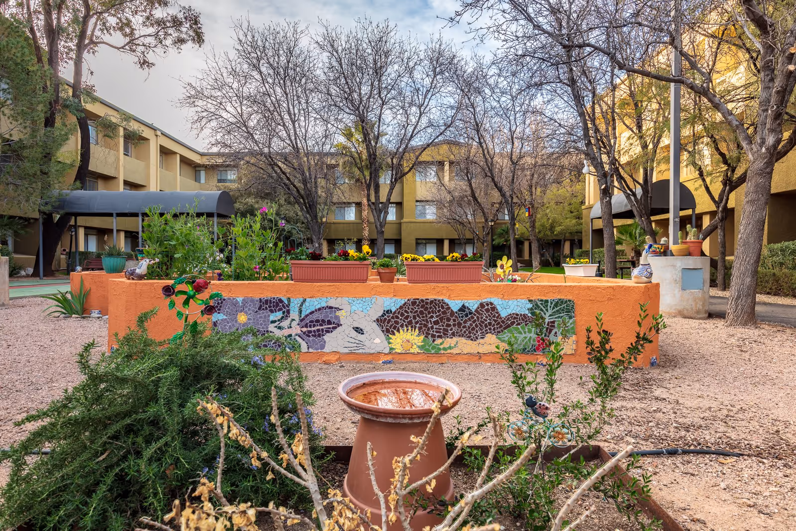 Outdoor courtyard area of Broadway Proper Assisted & Senior Living featuring a raised garden bed with colorful mosaic artwork on the side, various potted plants, leafless trees, and a multi-story building surrounding the courtyard.