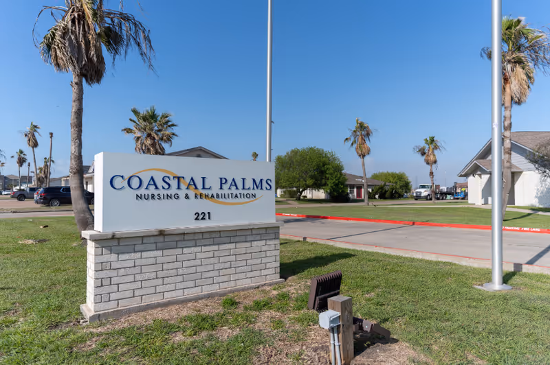 Outdoor view of the Coastal Palms Nursing & Rehabilitation facility sign with palm trees, grass, and buildings in the background under a clear blue sky.