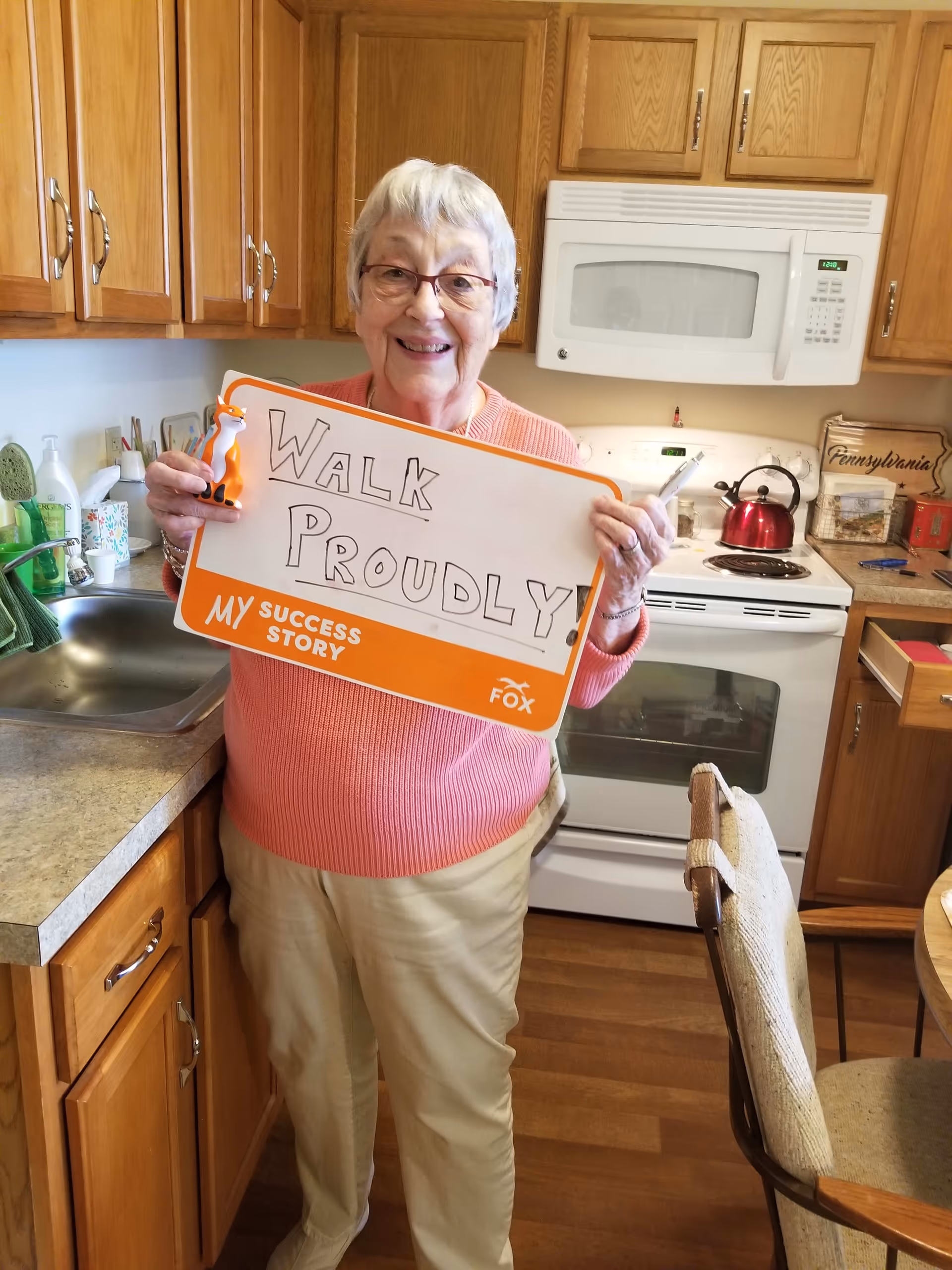 An elderly woman with short gray hair and glasses stands in a kitchen holding a sign that reads 'WALK PROUDLY!' She is smiling and wearing a pink sweater and beige pants. The kitchen has wooden cabinets, a white microwave, a white stove with a red kettle on it, and a sink with various items around it.
