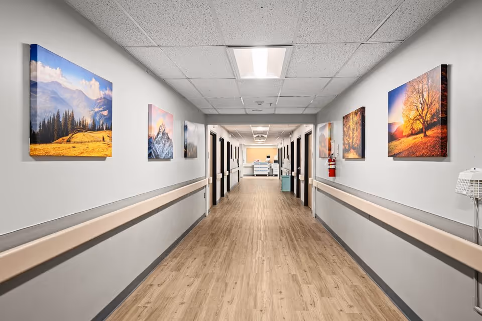 A long, well-lit hallway in a senior living facility with light wood flooring and white walls. The walls are decorated with colorful landscape paintings. Handrails run along both sides of the hallway, and several doorways are visible. At the end of the hallway, there is a reception or nurse station area.