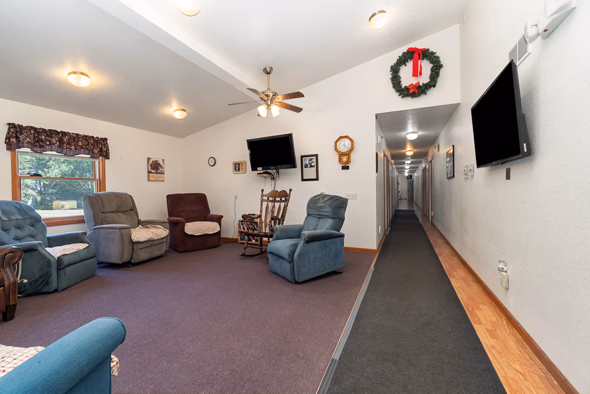 A senior living common room with several recliner chairs and a rocking chair facing wall-mounted TVs, a ceiling fan, and a long hallway to the right.