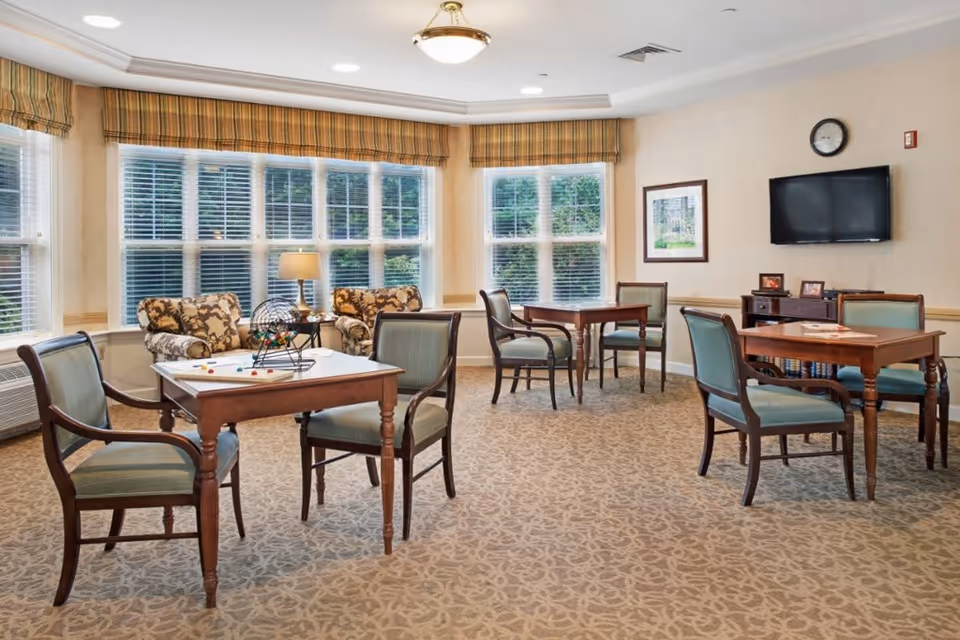 A bright and cozy common area with several wooden tables and chairs arranged for games or socializing. Two armchairs with floral upholstery are placed near large windows with blinds and striped valances. A table lamp sits on a small side table between the armchairs. On one table, there is a bingo cage and bingo balls. A flat-screen TV is mounted on the wall above a small cabinet with framed photos and books. The room has beige walls, a patterned carpet, and a ceiling light fixture.
