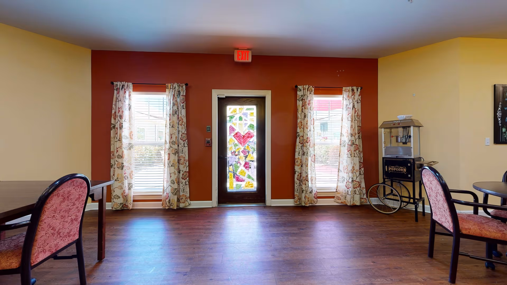 A cozy room with wooden flooring and warm-colored walls featuring a door with colorful stained glass in the center, flanked by two windows with floral curtains. There are several chairs with pink patterned upholstery and a popcorn machine on wheels to the right side of the room.