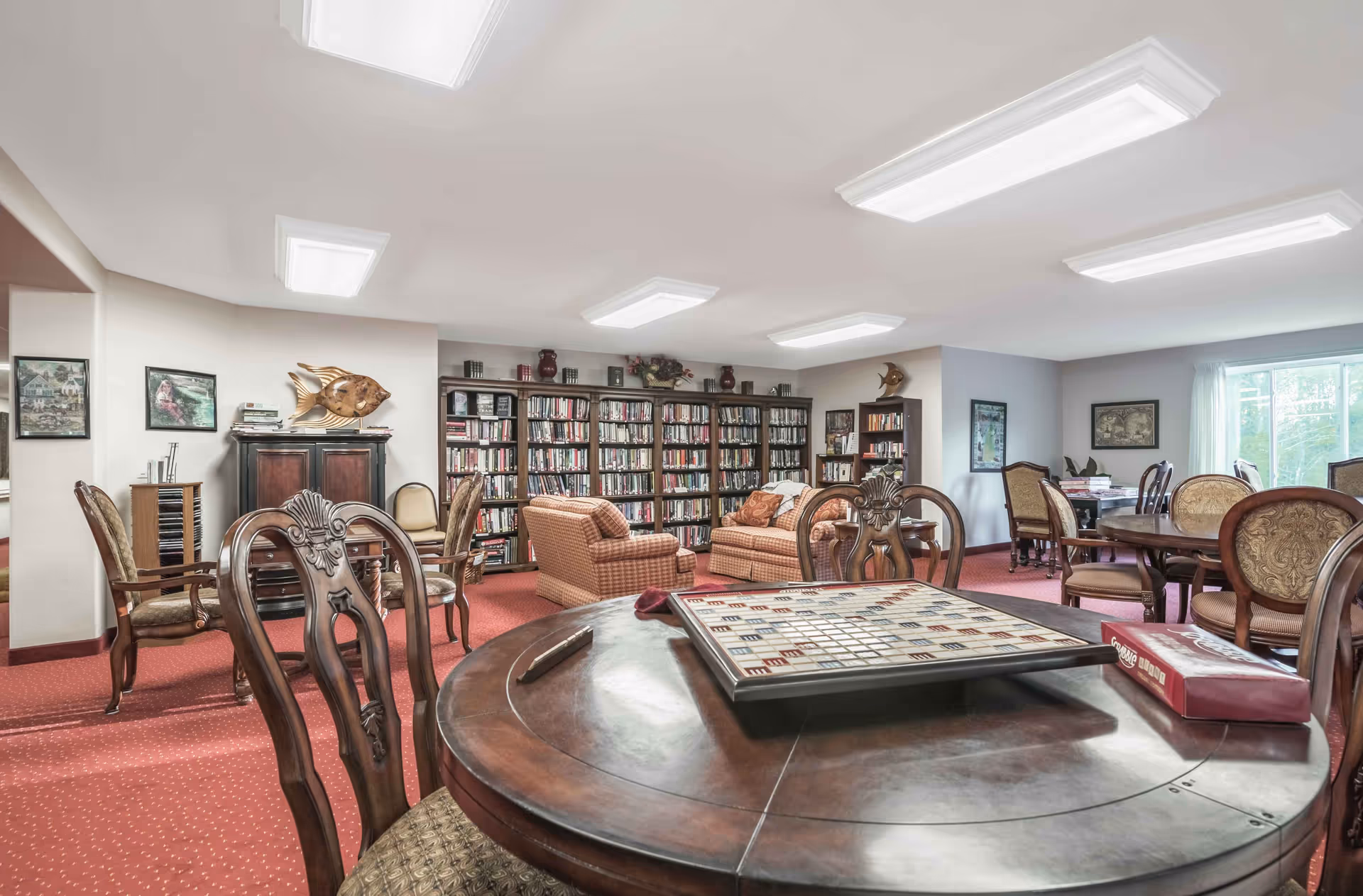 A bright communal library and game room with a round table holding a Scrabble board, upholstered chairs and sofas, bookshelves along the back wall, and large overhead lights.
