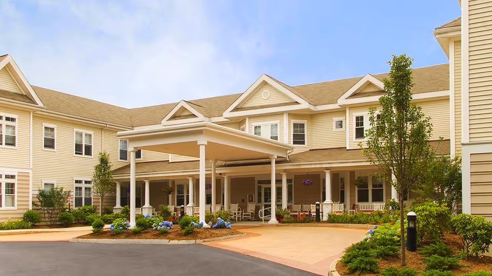 Exterior view of Atria Longmeadow Place, showing a two-story beige building with white trim, a covered entrance supported by columns, landscaped greenery, and a driveway.
