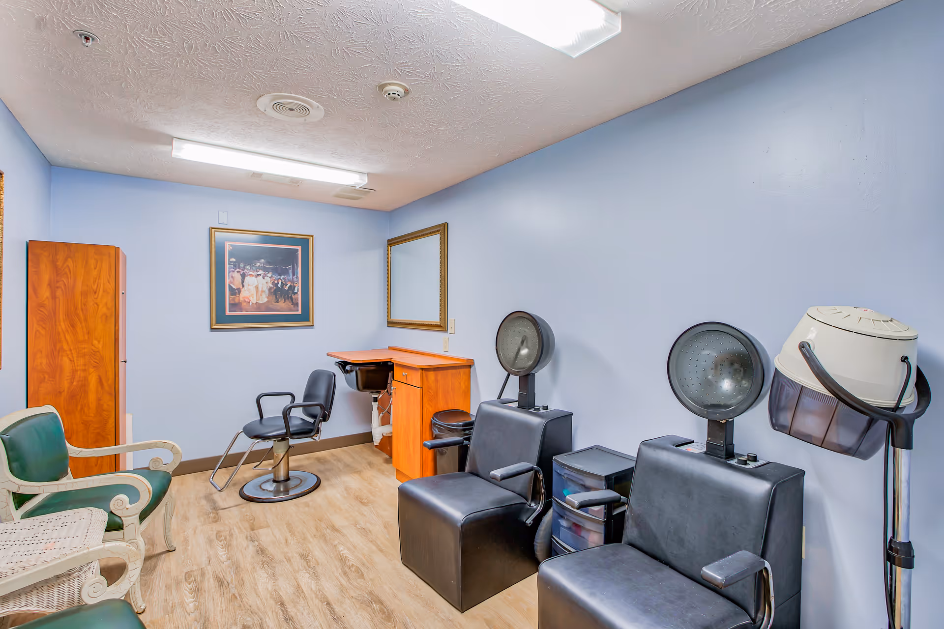 Interior view of a small salon room with light blue walls and wooden flooring. The room contains two black salon chairs with attached hair dryers, a black styling chair in front of a wooden counter with a mirror above it, and two green and white chairs along the left wall. A framed picture hangs on the back wall.