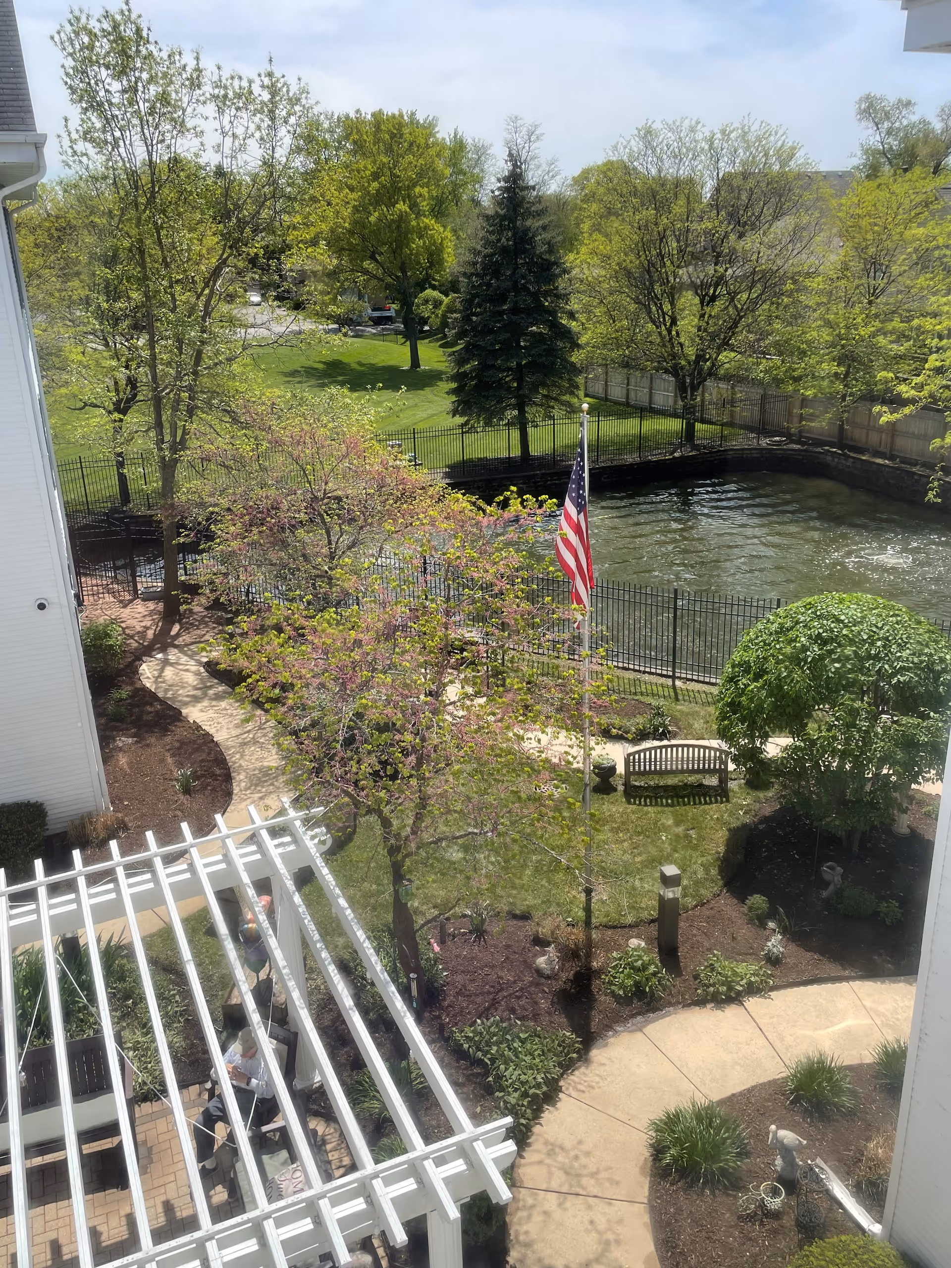 View of a landscaped outdoor garden area with a winding paved pathway, green trees, shrubs, and a small pond surrounded by a black metal fence. An American flag is displayed on a flagpole near a wooden bench. There is a white pergola structure with seating underneath, and a few people are visible sitting and standing in the shaded area.
