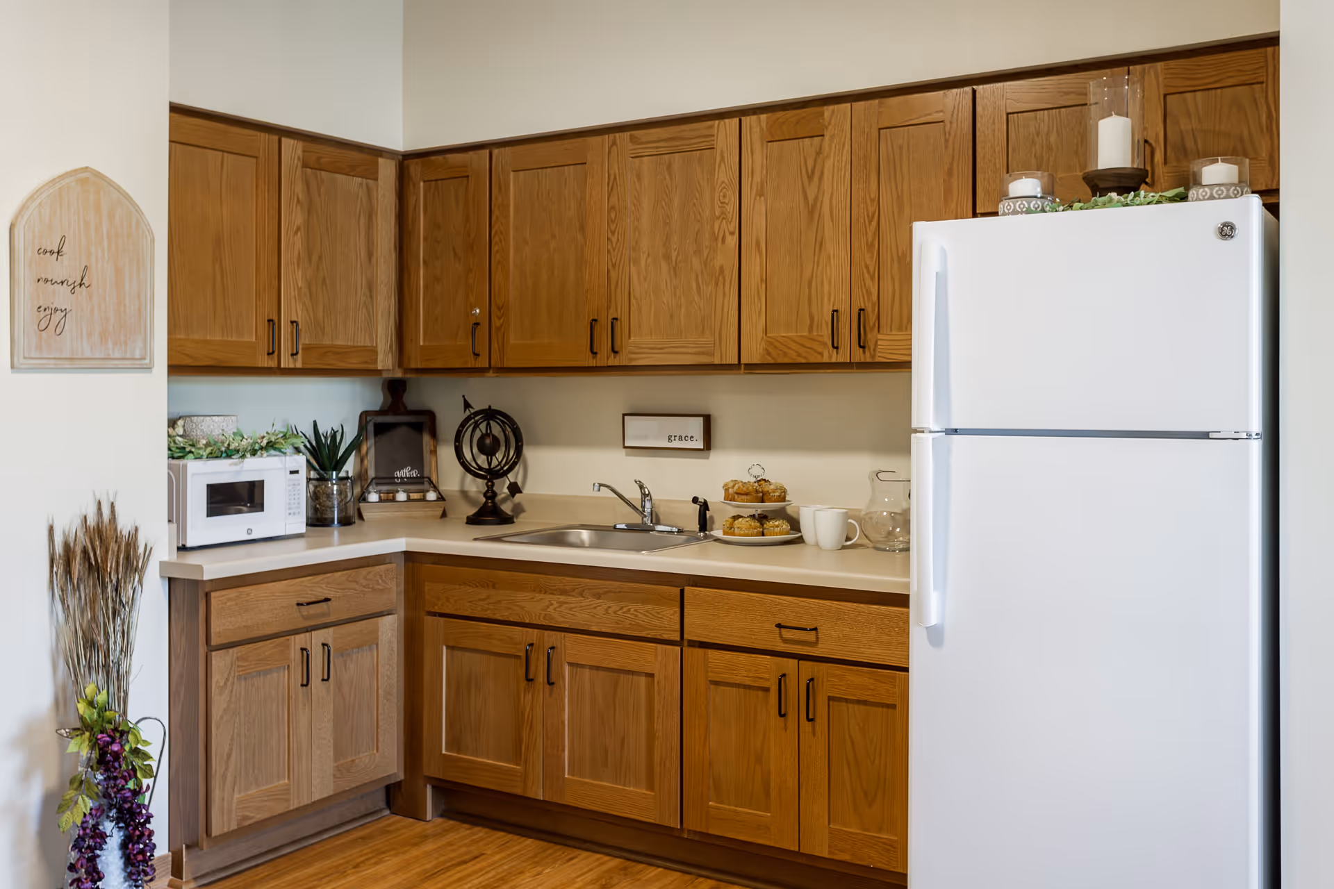 A kitchen area with wooden cabinets, a white refrigerator, a microwave, a sink, and various decorative items including candles, a small globe, and a plate of muffins. There is a wall decoration with the words 'cool, mindful, enjoy' and a bundle of dried plants in the corner.