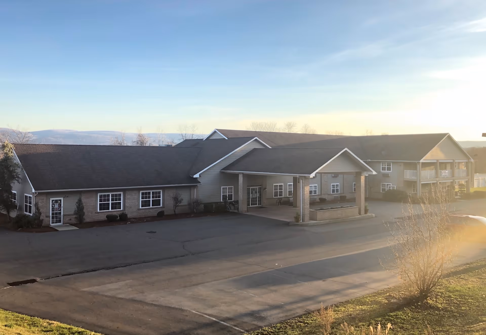 Exterior view of a single-story senior living facility building with a covered entrance and multiple windows, set against a backdrop of distant hills under a clear sky during sunset.