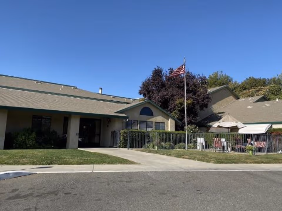 Exterior view of a single-story building with a sloped roof, an American flag on a flagpole, and a fenced patio area with outdoor furniture and an umbrella. The sky is clear and blue.