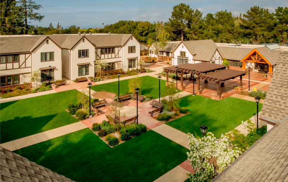 Aerial view of a senior living community courtyard with green lawns, paved walkways, benches, lamp posts, and pergolas. Surrounding the courtyard are two-story buildings with beige walls and brown trim, set against a backdrop of trees and a clear sky.