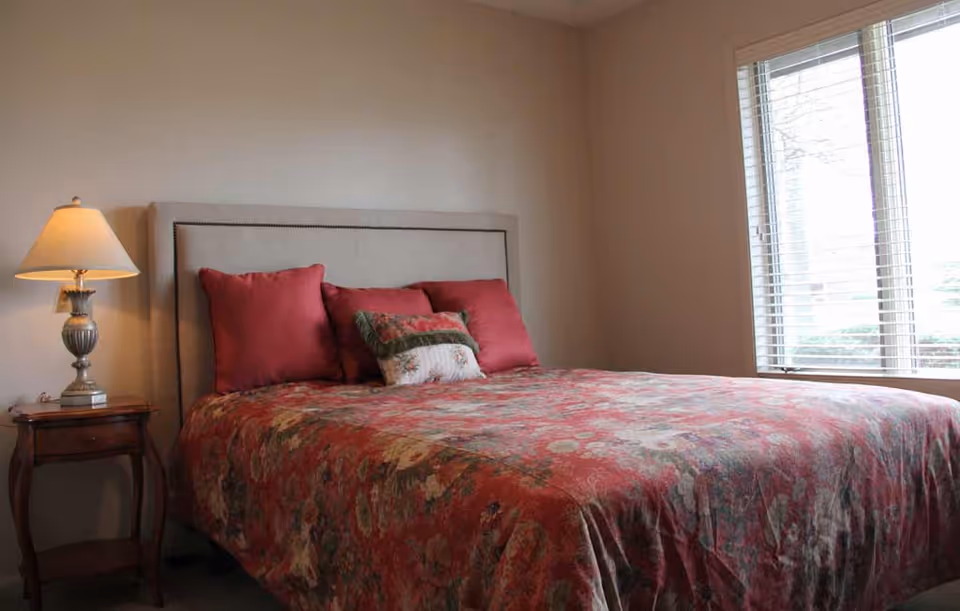 A bedroom with a bed covered in a floral patterned red bedspread and several pillows. There is a wooden nightstand with a lamp on it to the left of the bed, and a window with blinds on the right side letting in natural light.