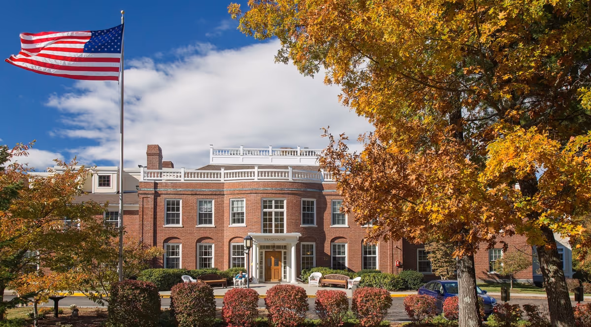 Brick colonial-style senior living building entrance with an American flag and fall foliage in front.