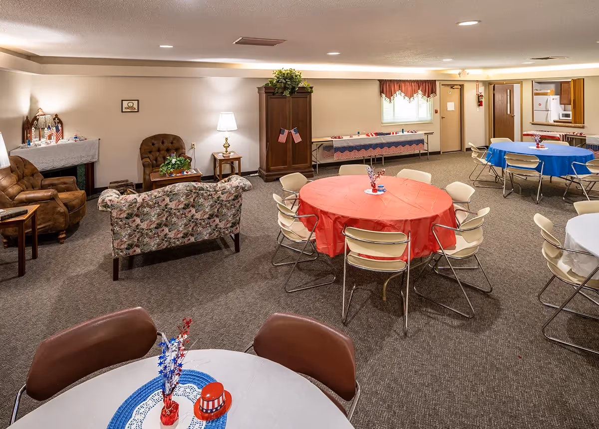 A common area in a senior living facility with multiple round tables covered with red, blue, and white tablecloths and decorated with patriotic centerpieces. There are several chairs around the tables. In the background, there is a seating area with a floral couch, two brown armchairs, a wooden cabinet, and a side table with a lamp. The room has carpeted floors and neutral-colored walls.