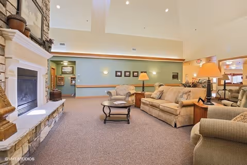 A spacious living room area in a senior living facility featuring a stone fireplace on the left, beige sofas and armchairs arranged around a wooden coffee table, soft lighting from table lamps, and framed pictures on a green accent wall in the background.