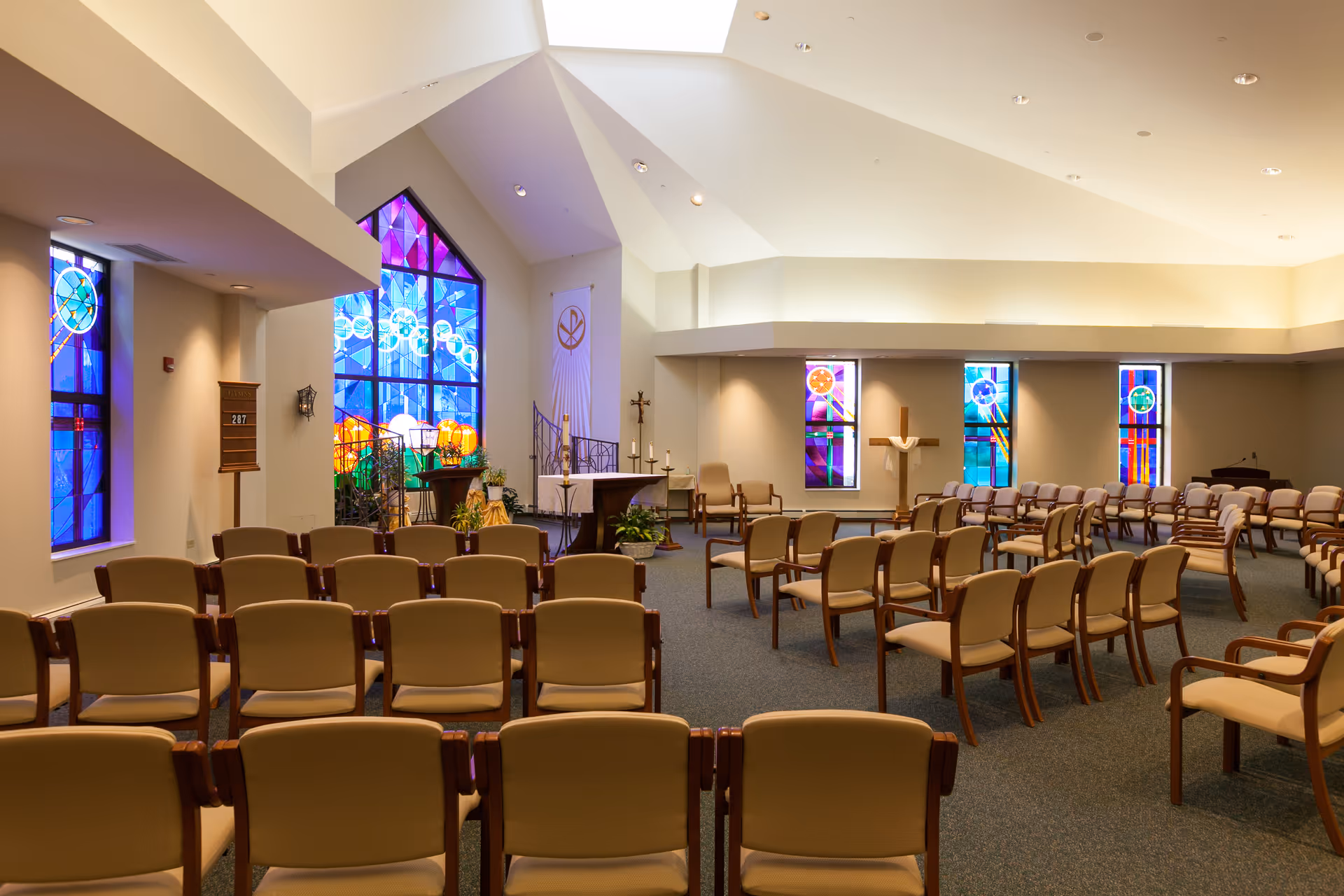 Interior view of a chapel with rows of beige chairs facing an altar. The chapel features colorful stained glass windows, a wooden cross, and a high vaulted ceiling with recessed lighting.