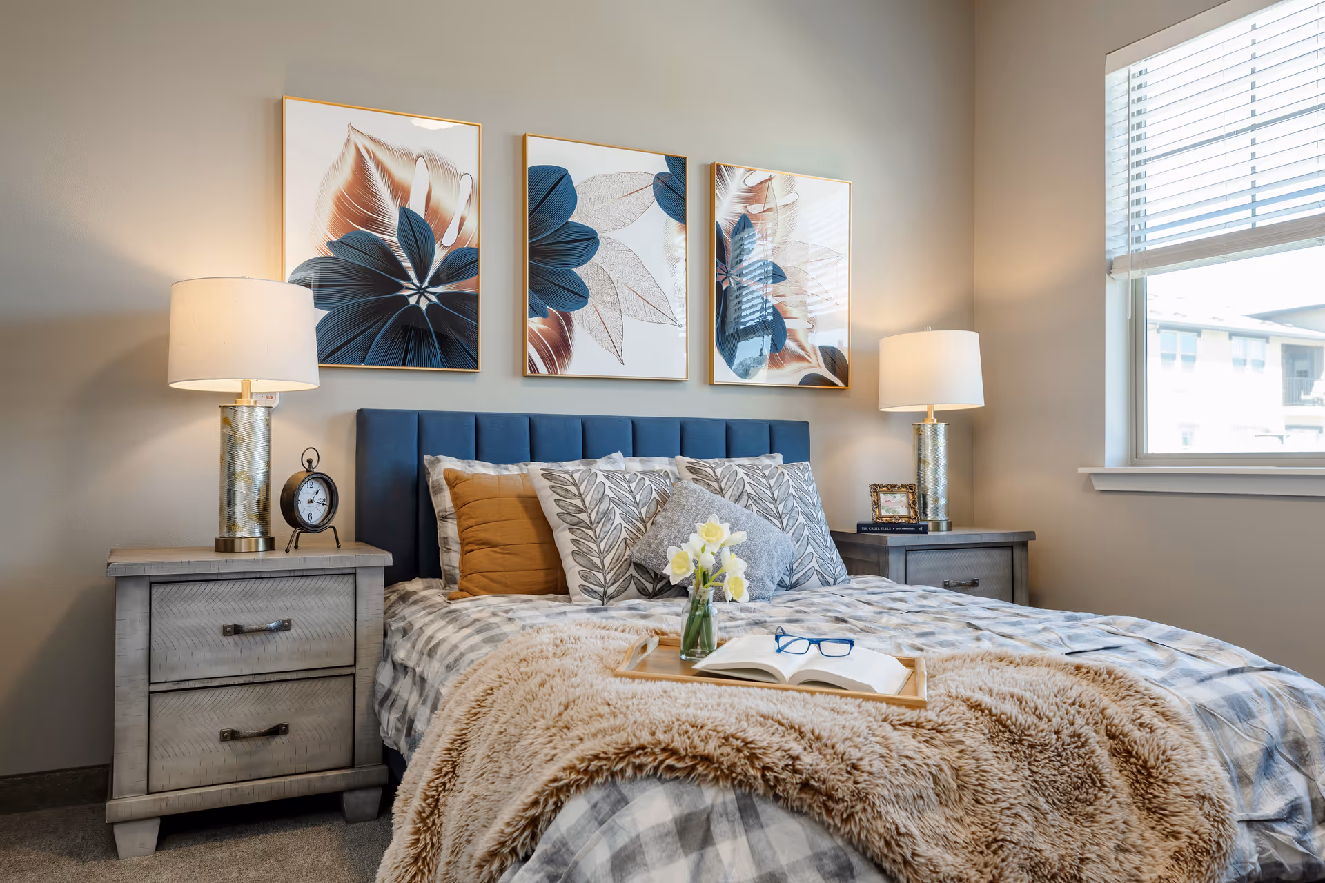 A cozy bedroom featuring a bed with a blue upholstered headboard, plaid bedding, decorative pillows, and a soft brown throw blanket. On the bed is a wooden tray holding a vase with yellow flowers, an open book, and a pair of glasses. Two matching nightstands flank the bed, each with a lamp and decorative items. Above the bed are three framed floral artworks. A window with blinds allows natural light into the room.