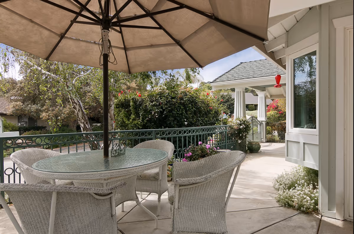 Outdoor patio area with a round glass-top table surrounded by four white wicker chairs under a large beige umbrella. The patio is bordered by a green metal railing and surrounded by lush greenery and flowering plants. A covered porch with white columns and a hanging red bird feeder is visible in the background.