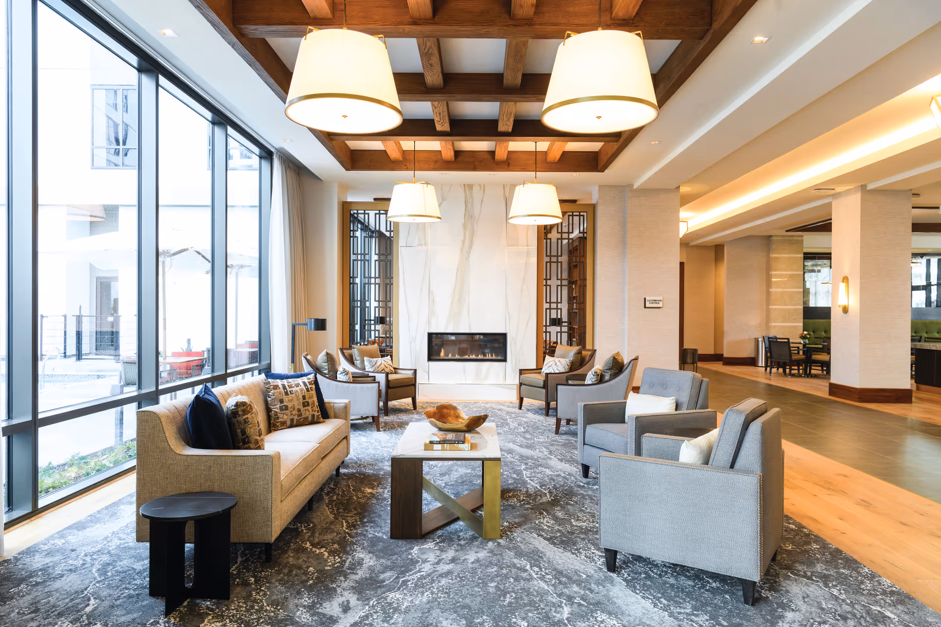 A spacious and modern living room area with large floor-to-ceiling windows on the left, allowing natural light to fill the space. The room features a beige sofa with decorative pillows, several gray armchairs, a central coffee table with a decorative bowl, and a contemporary fireplace set in a white marble wall. The ceiling has wooden beams with large hanging light fixtures, and the floor is covered with a patterned gray rug. In the background, there is an open area with more seating and wooden flooring.