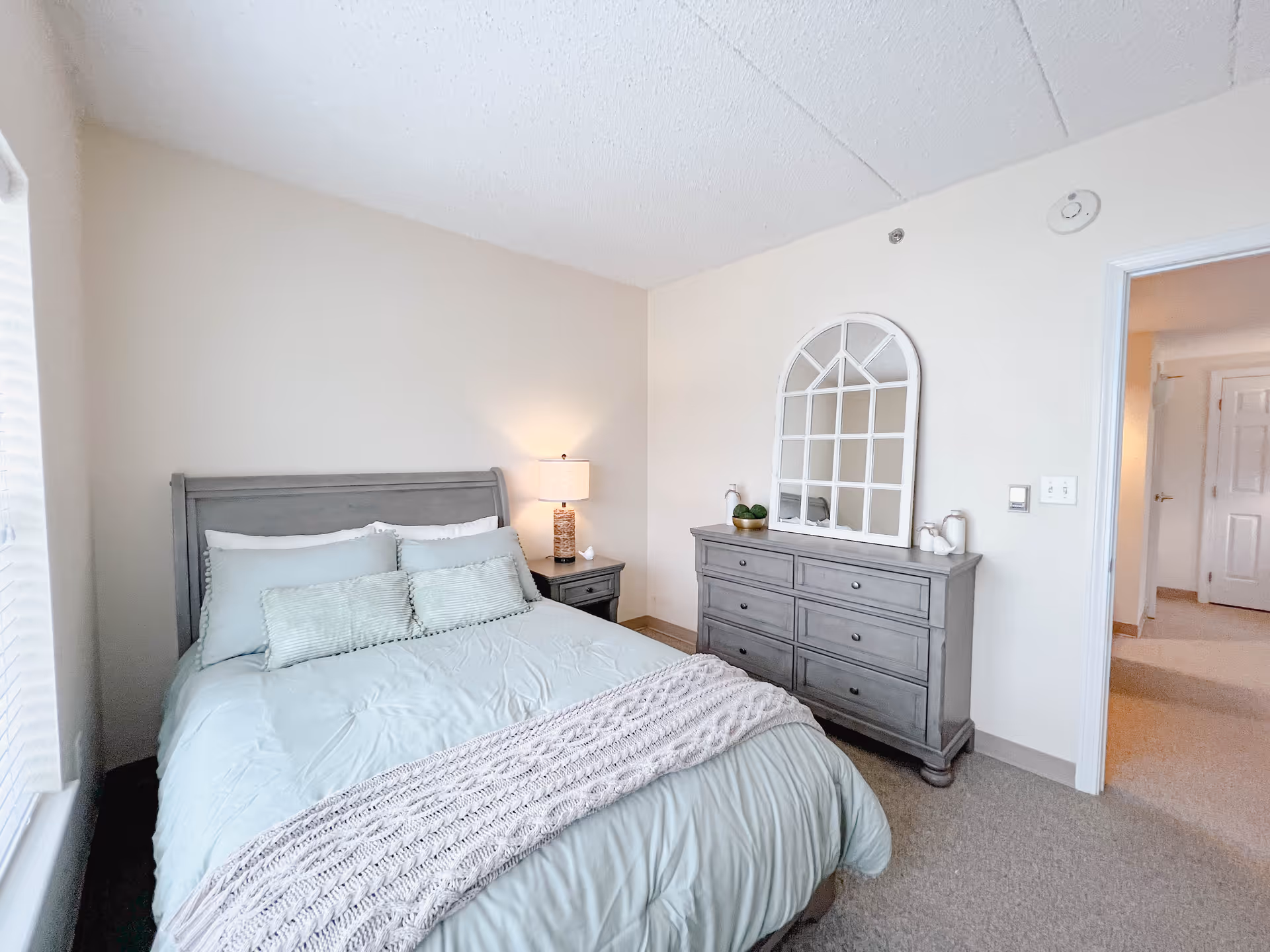 A tidy bedroom with a gray wooden bed dressed in light blue bedding, a nightstand with a lamp, and a matching dresser topped by an arched mirror.